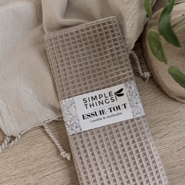 A beige waffle-patterned towel titled "Seasoned" from Sonia's Vokalized Kreations elegantly rests on a light wooden surface with a matching cloth and lush green plant, evoking the vibrant flavors of a Barbados hot pepper sauce.