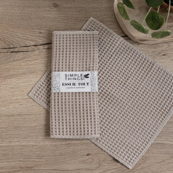 Two beige waffle-textured kitchen towels, one labeled "Simple Things," rest on a wooden surface. Nearby, a small plant in a ceramic dish and the "Seasoned" Afro-Vegan cookbook from Sonia's Vokalized Kreations await culinary adventures.