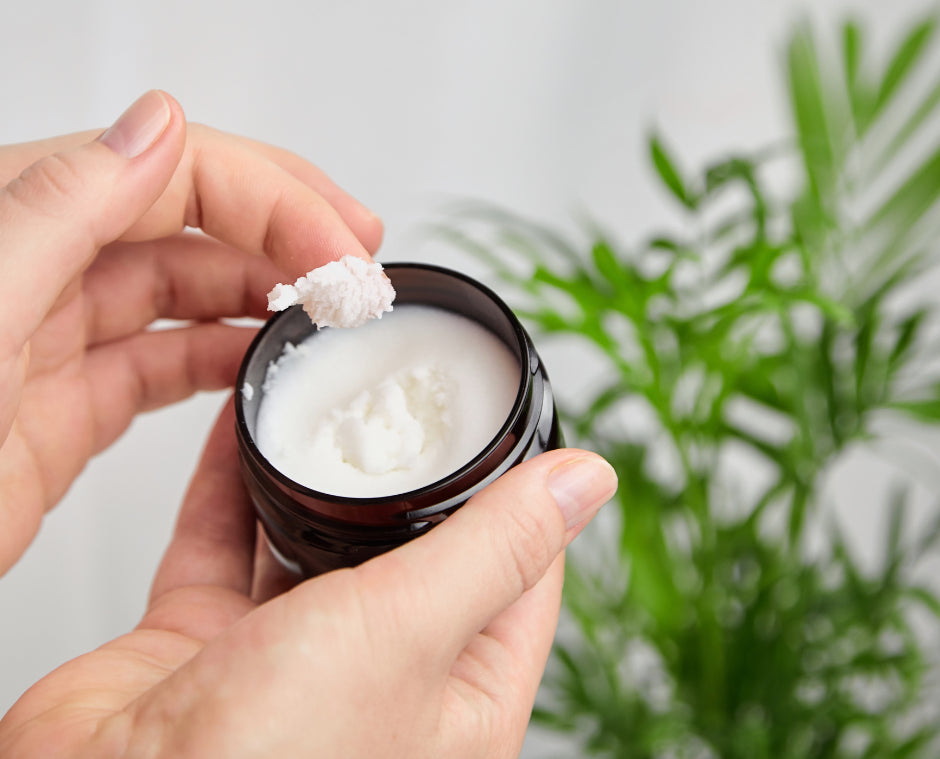 A person scoops white balm from a dark jar with a green plant nearby—perfect for thoughtful postpartum care or as part of the Sonia's Vokalized Kreations New Baby Gift Box for Mom & Baby, a custom shower or postpartum bundle.