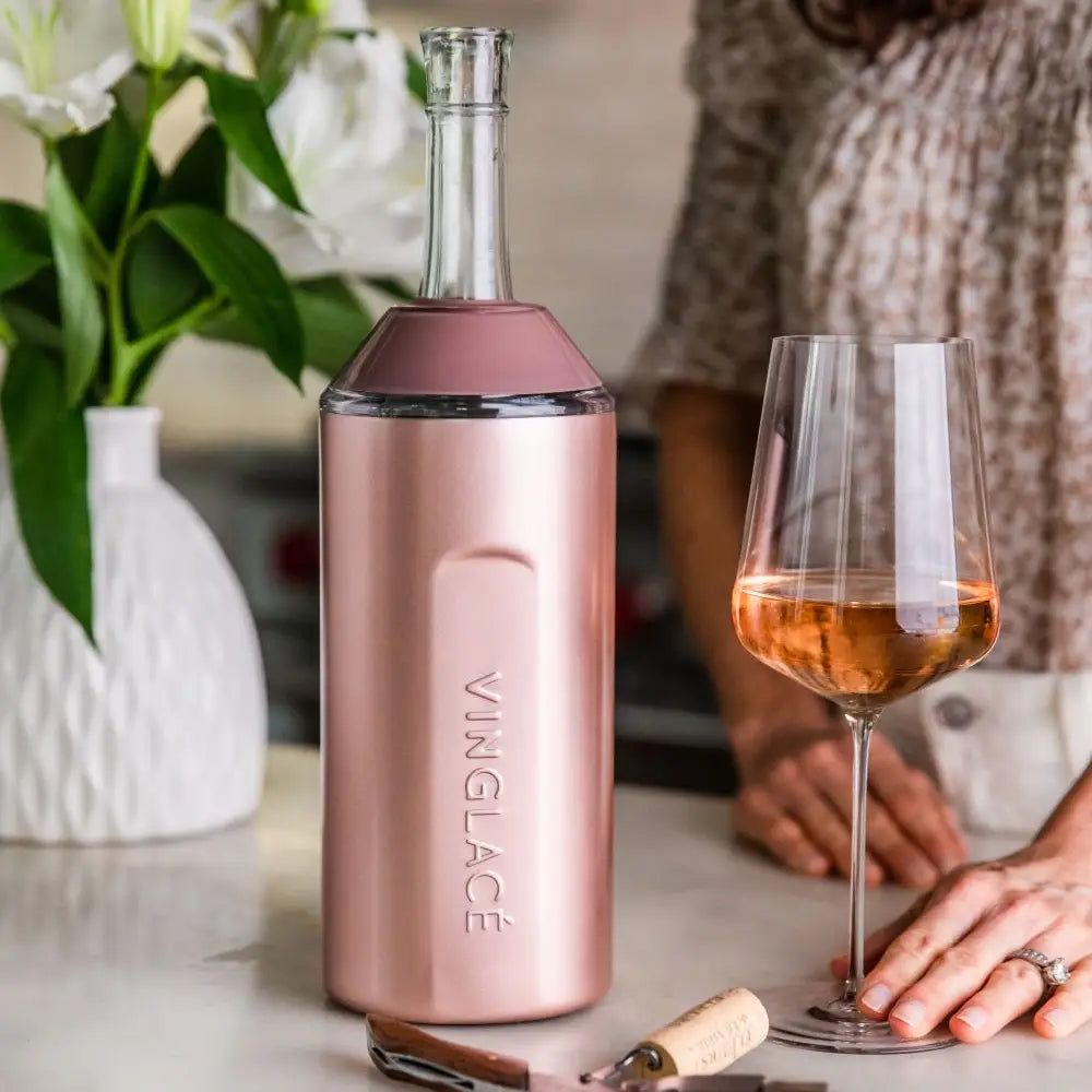 A Wine & Champagne Chiller from sonia's-vokalized-kreations, in rose gold, holds a bottle on the table beside a filled glass. Nearby, someone admires the scene decorated with flowers and a cork.