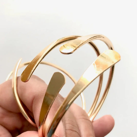 A hand displays Brass Coil Bangles by Sonia's Vokalized Kreations—modern, shiny, gold-colored open-ended metal bangles set against a white background.