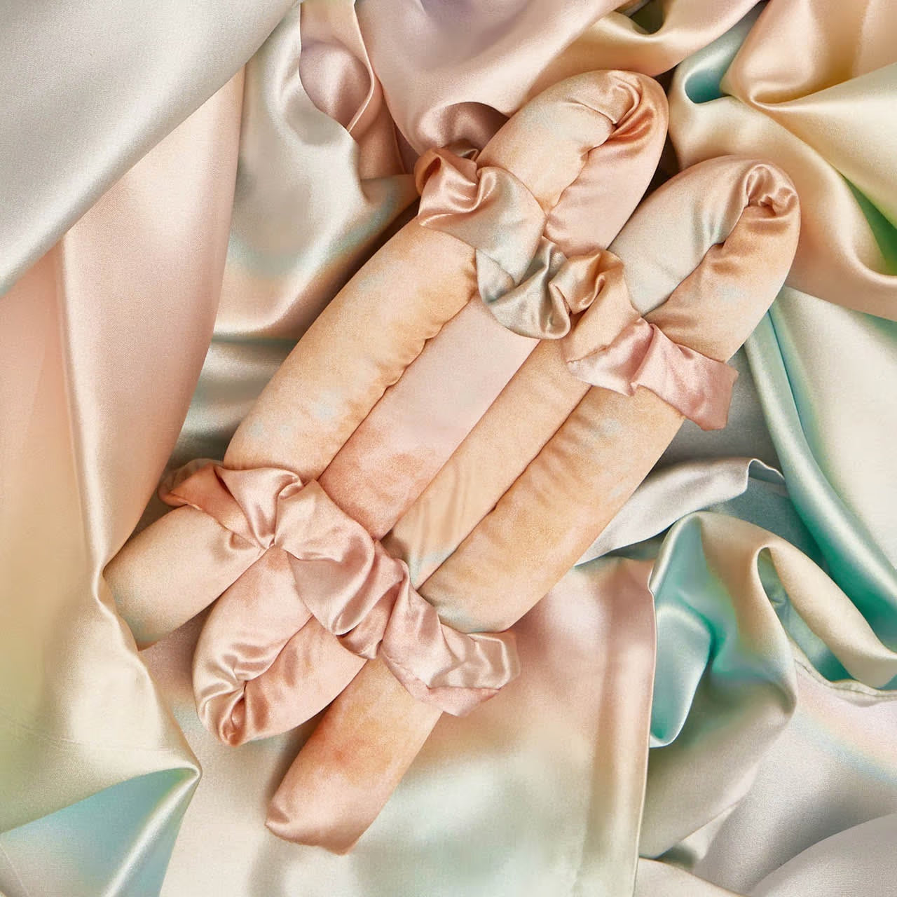 Silk-covered Self Less Care hair rollers from Sonia's Vokalized Kreations, in pastel shades, rest on crumpled, satin-like fabric of soothing hues, inviting a moment of tranquility and self-care.