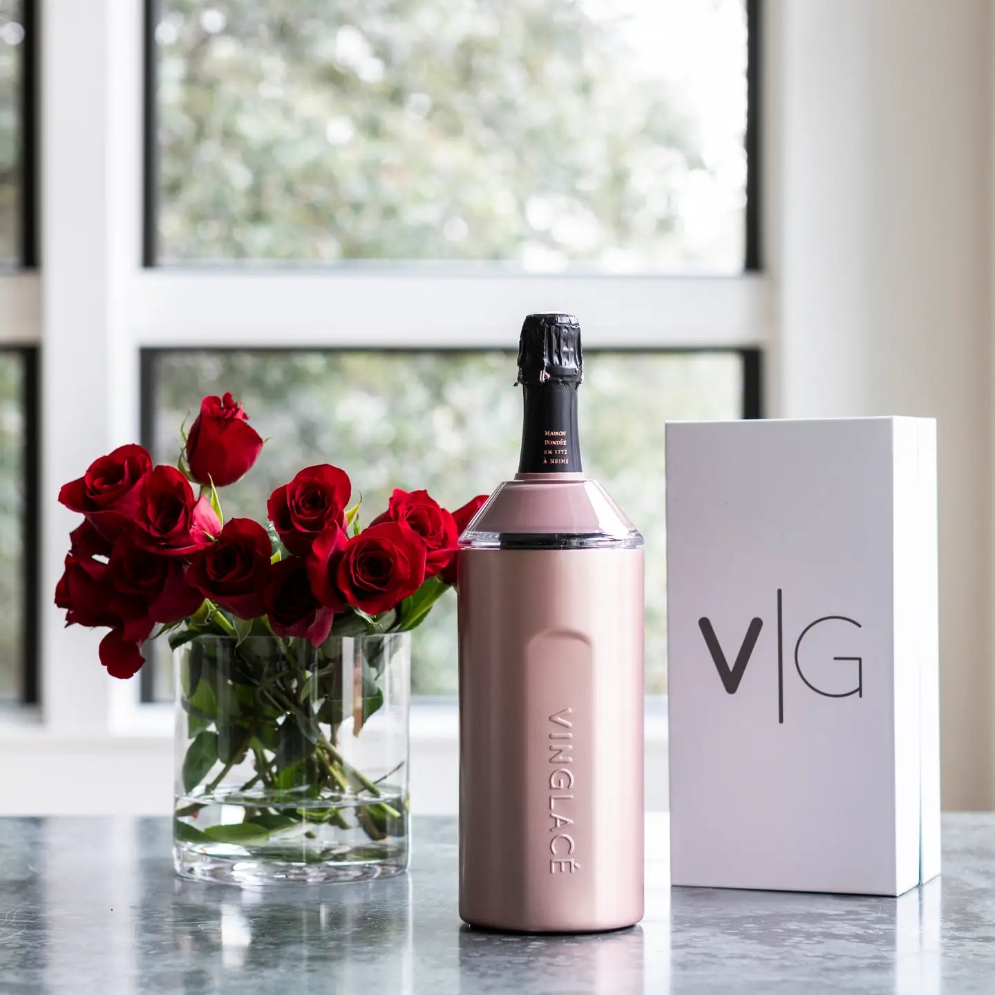 A rose gold Wine & Champagne Chiller by sonia's-vokalized-kreations and a sleek white box sit elegantly beside a vase of red roses on the table, illuminated by sunlight through the window.