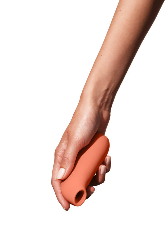 A hand holds the Aer Suction Toy, an orange cylindrical object from Sonia's Vokalized Kreations, made of medical-grade silicone against a white background.