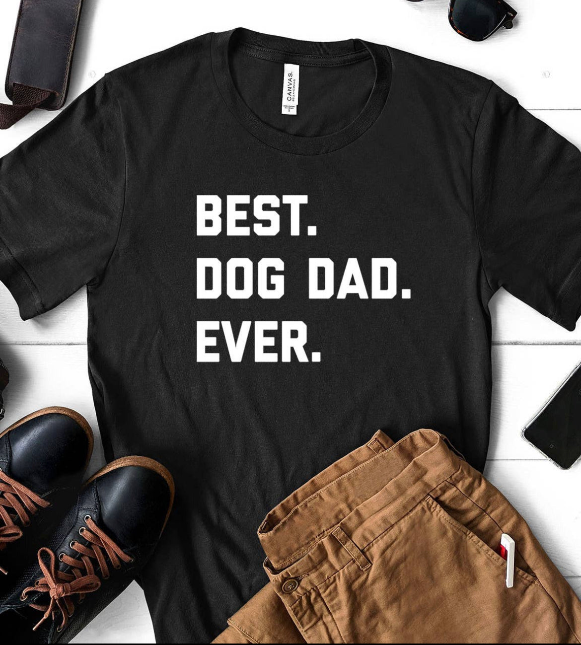 The "Best Dog Dad Ever" T-shirt by sonia's-vokalized-kreations is a unisex 100% cotton piece, paired stylishly with brown pants, sunglasses, and shoes on a white wooden background.