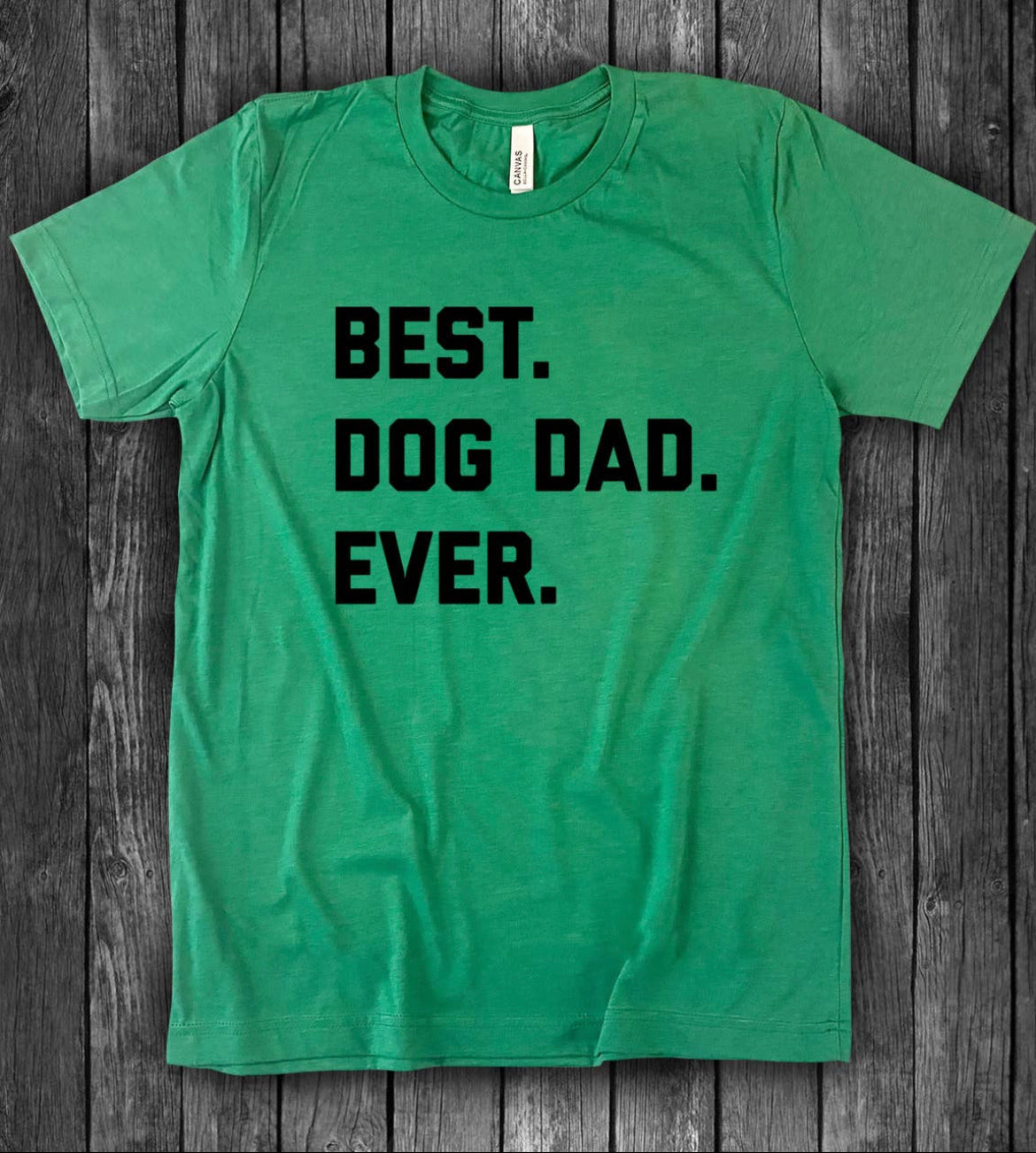 The Best Dog Dad Ever T-Shirt by sonia's-vokalized-kreations is a unisex green tee with bold black text on a wooden backdrop, crafted from 100% cotton for ultimate comfort.