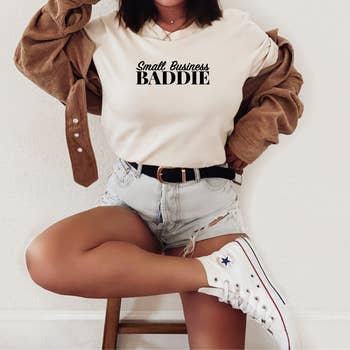 A person seated, sporting a "Small Business Baddie T-Shirt" by Sonia's Vokalized Kreations, paired with a brown jacket, ripped denim shorts, and white high-top sneakers—truly championing small businesses.