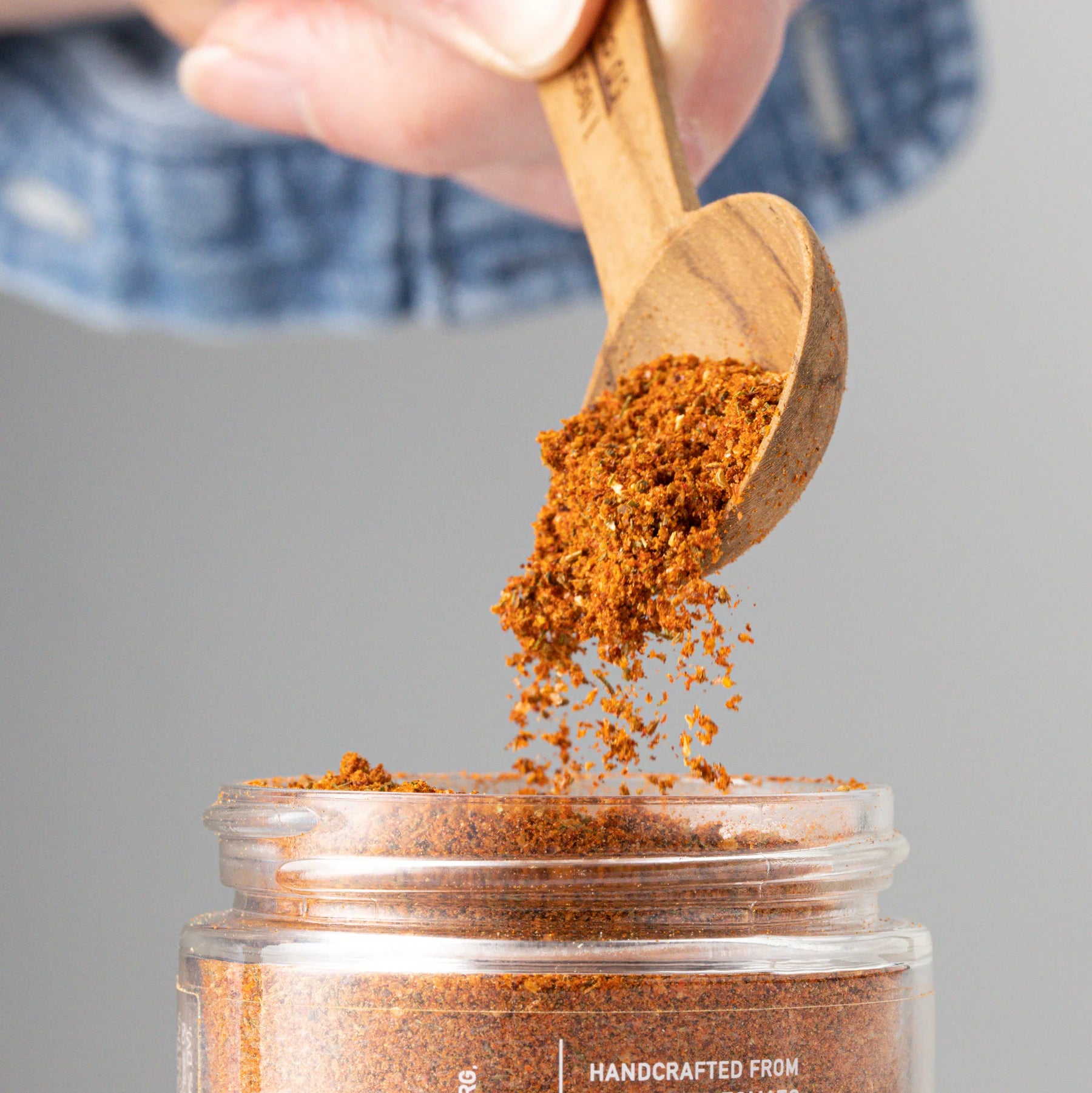 A hand delicately sprinkles Seasoned, a seafood spice blend by Sonia's Vokalized Kreations, into a jar labeled handcrafted, set against a blurred neutral background that captures culinary artistry.