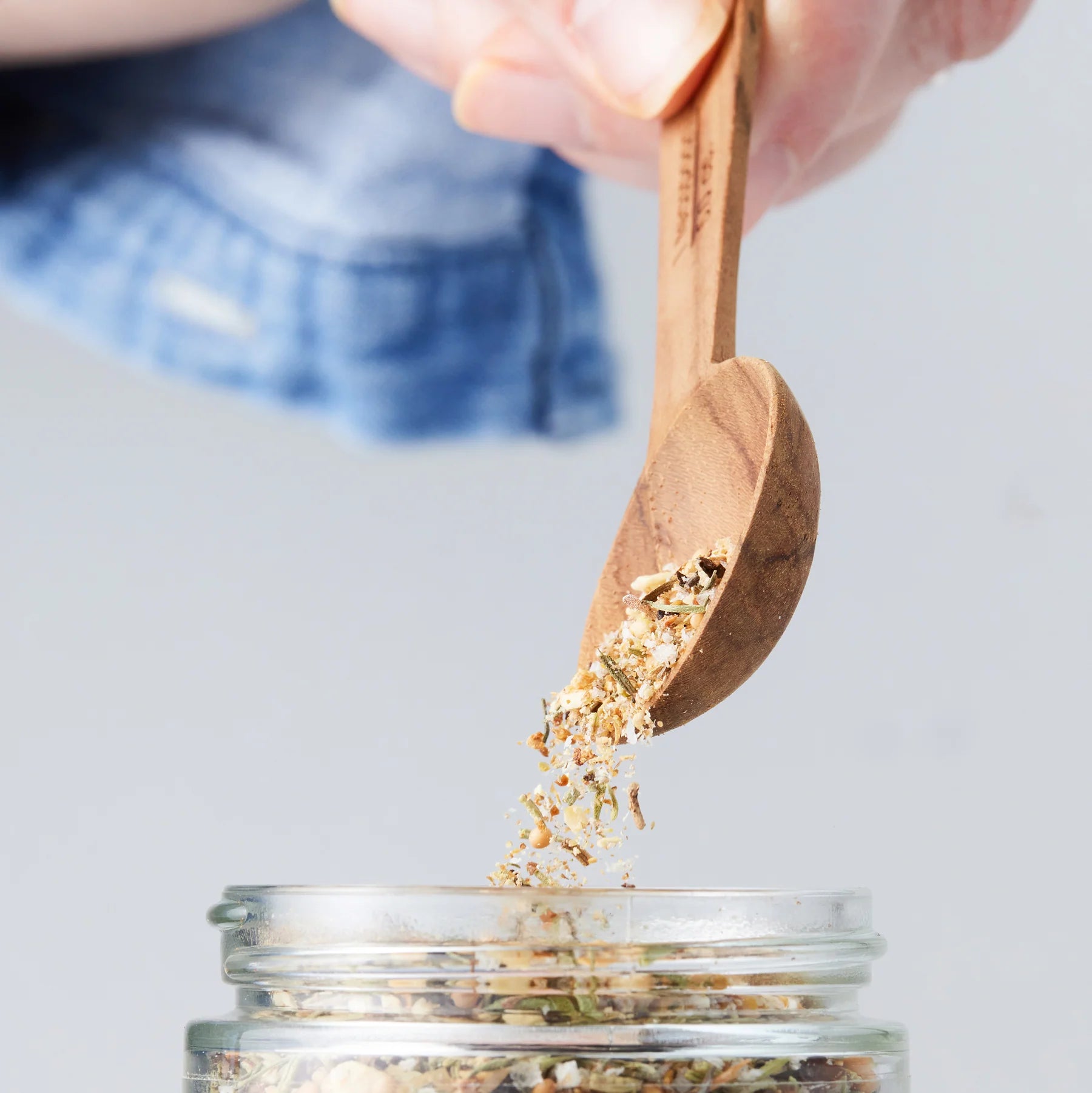 A hand uses a wooden spoon to pour herbs from "Seasoned," an Afro-Vegan cookbook by Sonia's Vokalized Kreations, into a glass jar.