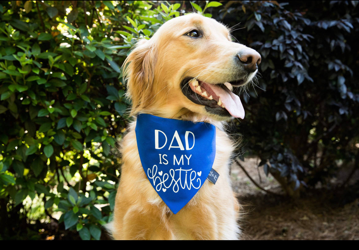 A stylish golden retriever, wearing a sonia's-vokalized-kreations Dog Bandana with "Dad is my bestie" on it, poses adorably against lush green foliage.