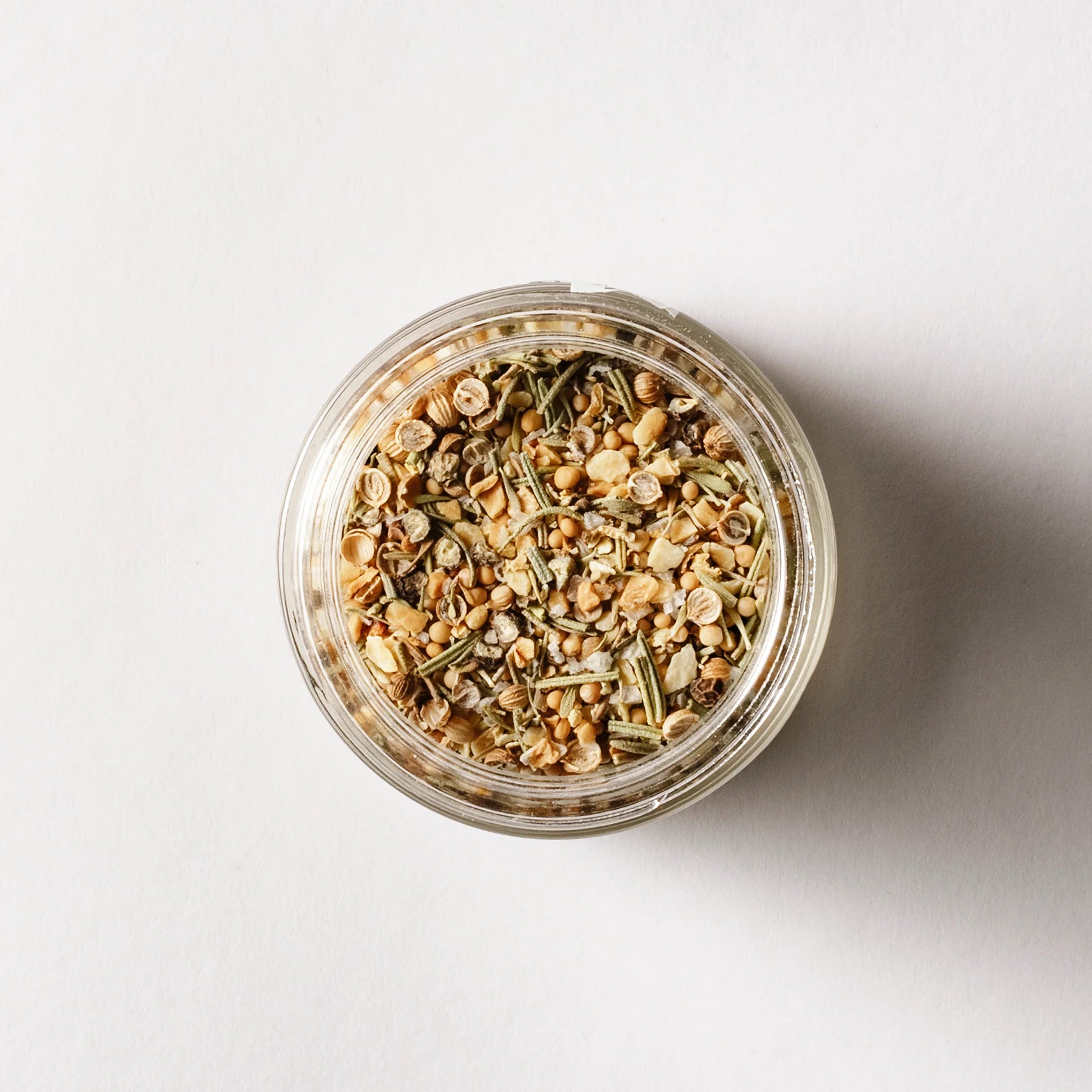 An overhead view shows a glass jar of "Seasoned" by Sonia's Vokalized Kreations, containing a vibrant seafood spice blend with an enticing mix of dried herbs and seeds, set against a plain background.