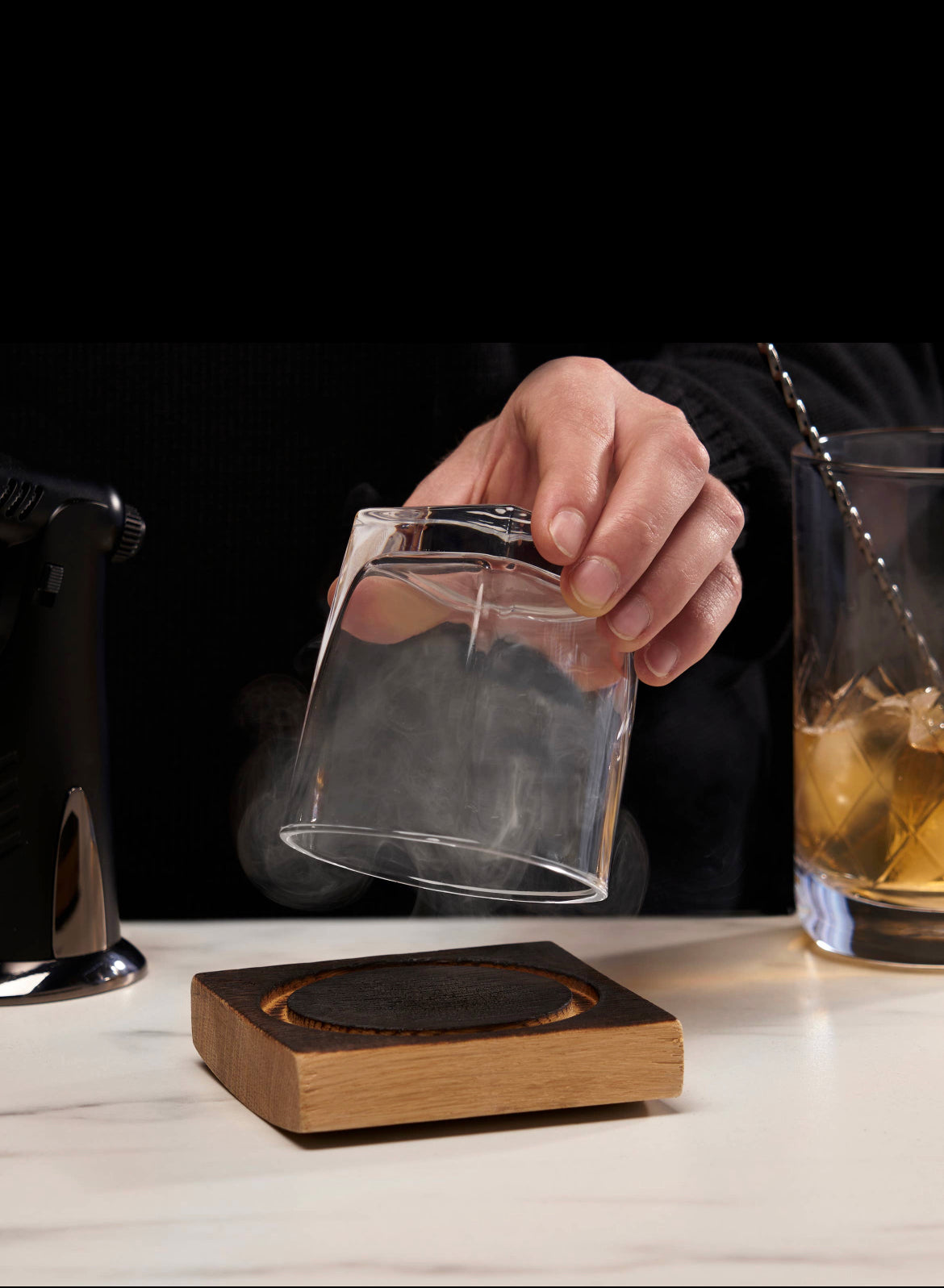 A hand holds an upside-down glass capturing smoke from a wooden coaster, thanks to the AlchemI Single Serve Smoked Cocktail Kit by sonia's-vokalized-kreations, with a cocktail glass and mixer gracefully set in the background.