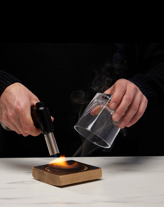 Someone is preparing their AlchemI Single Serve Smoked Cocktail Kit from sonia's-vokalized-kreations, using a handheld torch to light the wooden disc on a coaster, with a clear glass placed nearby on a white surface.