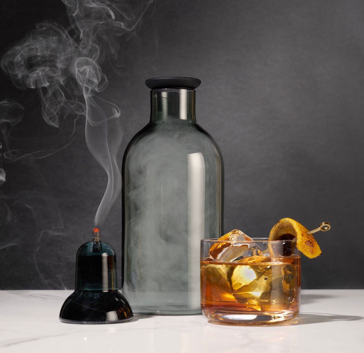 The Alchemi Smoked Cocktail Kit by sonia's-vokalized-kreations features a glass bottle with smoke beside a smoking cloche and an iced drink garnished with fruit on a stick. Set against a dark background, this scene suggests the smoke rinse method, enhancing the whiskey-soaked oak aroma.