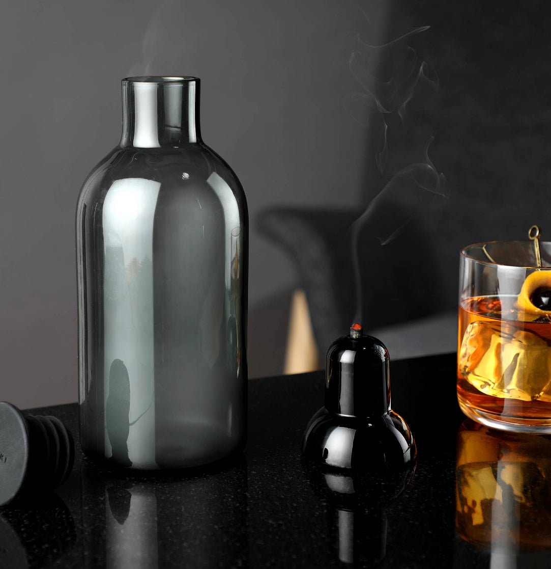 The Alchemi Smoked Cocktail Kit by sonia's-vokalized-kreations features a glass bottle, black incense holder releasing smoke, and a glass of amber liquid with lemon slices and an olive on a glossy black surface, evoking whiskey-soaked oak.