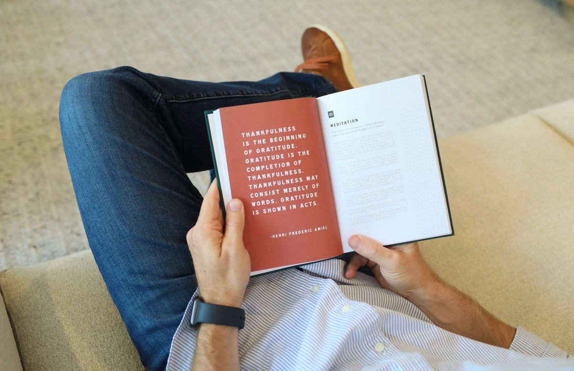 Immersed in a book with a quote on thankfulness, a person in blue jeans and a white shirt sits on a light-colored couch. Their smartwatch aids mindfulness routines as the page from "Men's Gratitude Journal" by sonia's-vokalized-kreations becomes their daily gratitude practice.