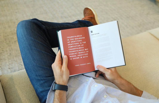 Immersed in a book with a quote on thankfulness, a person in blue jeans and a white shirt sits on a light-colored couch. Their smartwatch aids mindfulness routines as the page from "Men's Gratitude Journal" by sonia's-vokalized-kreations becomes their daily gratitude practice.