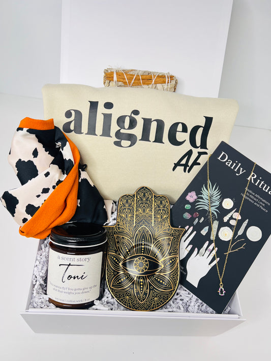 The "Don't Disturb My Peace" gift box by Sonia's Vokalized Kreations includes a fabric pouch with "aligned AF," a black and white scarf with orange accents, an "a scent story Toni" candle, hand-shaped decor, and a "Daily Rituals" booklet—each item inspiring inner harmony and mindfulness.