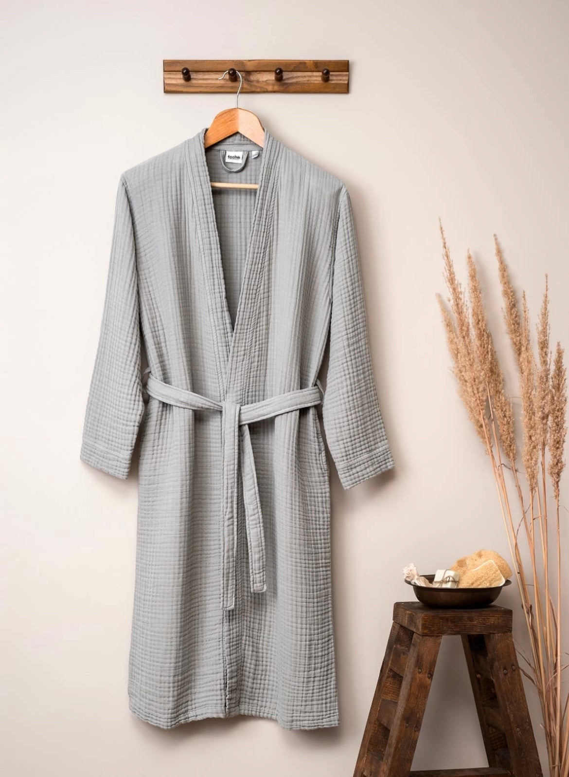 A Gauze Cotton Muslin Bathrobe by sonia's-vokalized-kreations, made in Turkey from 100% cotton, hangs on a wooden hook. A wooden stool nearby supports a bowl of natural sponges and a stone, with dried plants adding texture to the background.