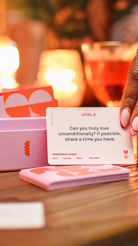 A hand holds a Touchy/Feely™ card from Sonia's Vokalized Kreations' intimacy game, asking, "Can you truly love unconditionally? If possible, share a time you have." Erogenous zones like chin and calves invite deeper connections between partners.