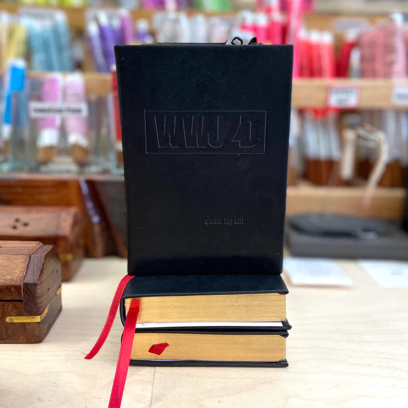 A black book embossed with "WWJD," featuring a red ribbon bookmark, sits on two gold-edged volumes. Shelves of colorful threads in the background showcase the artistry of svkreations by svkreations.