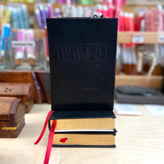 A black book embossed with "WWJD," featuring a red ribbon bookmark, sits on two gold-edged volumes. Shelves of colorful threads in the background showcase the artistry of svkreations by svkreations.