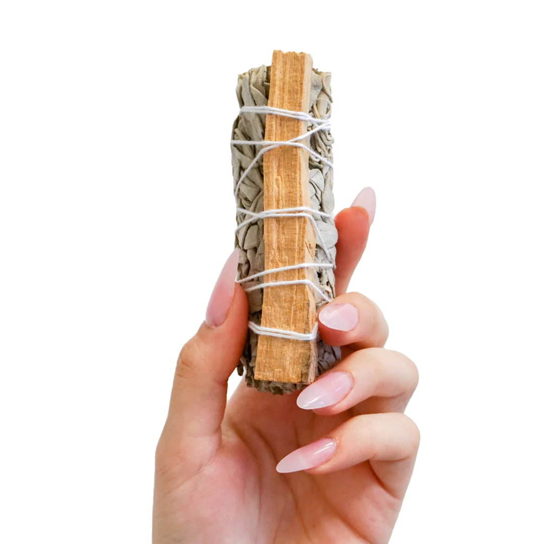 A hand delicately holds Sonia's Vokalized Kreations' "Don't Disturb My Peace," a bundle of sage and palo santo tied with white string against a white backdrop, symbolizing serenity and inviting mindfulness into daily life.