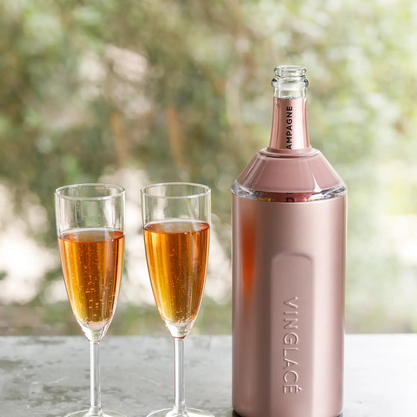 A bottle of champagne is in a sonia's-vokalized-kreations Wine & Champagne Chiller, flanked by two filled champagne flutes on a table, with a blurred green backdrop.