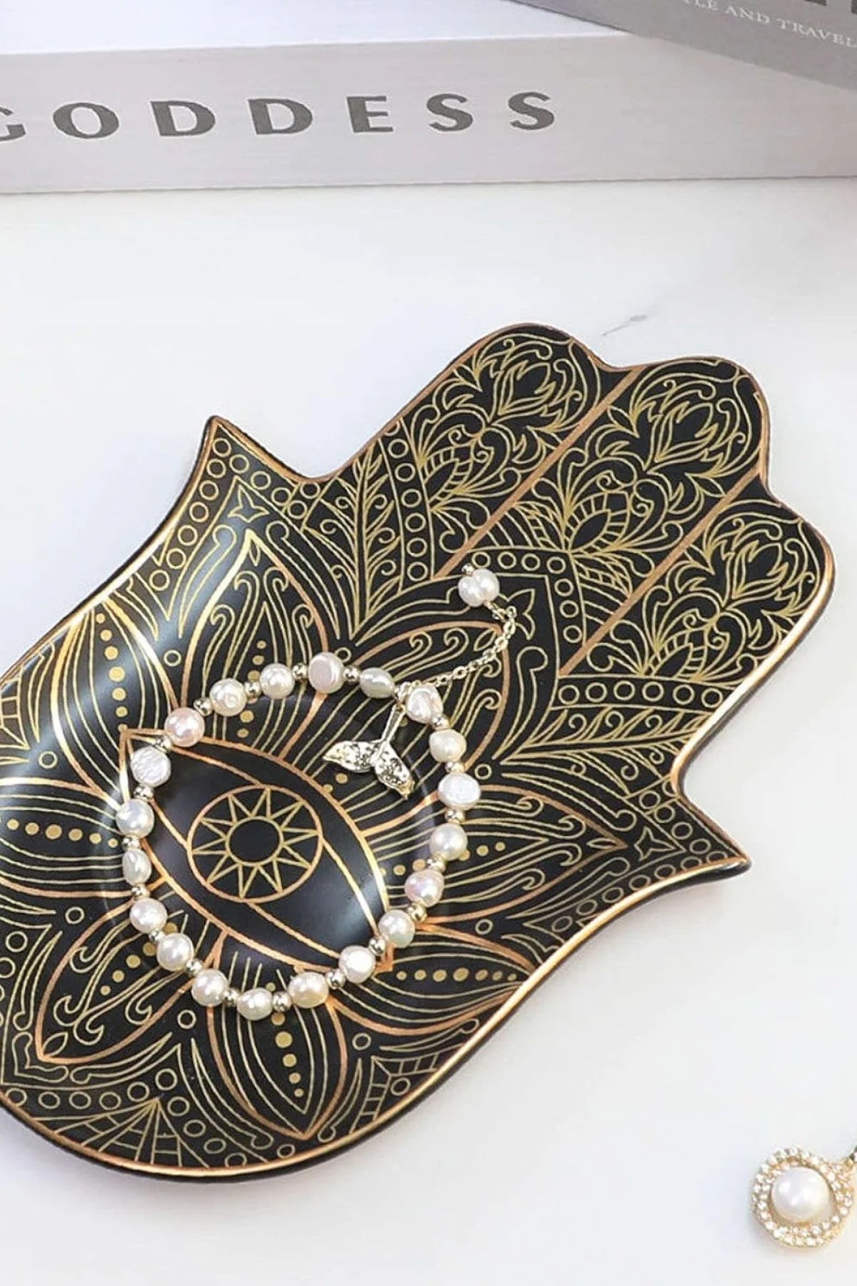 The "Don't Disturb My Peace" tray from Sonia's Vokalized Kreations, adorned with gold patterns and holding a pearl bracelet, exudes tranquility. A partial view of a pearl ring adds harmony, while the glimpse of a book titled "GODDESS" suggests mindfulness in every detail.