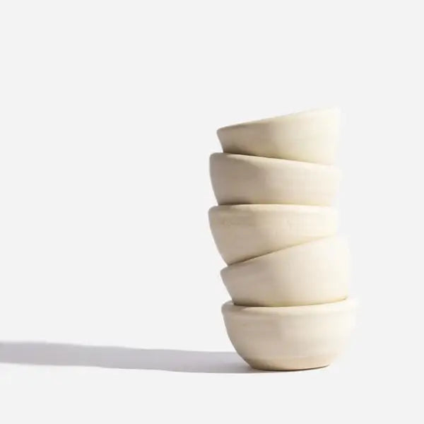 A set of five off-white ceramic bowls from Sonia's Vokalized Kreations, named the Mask Bowl & Brush Set, elegantly stacked in a slightly leaning tower against a plain background.