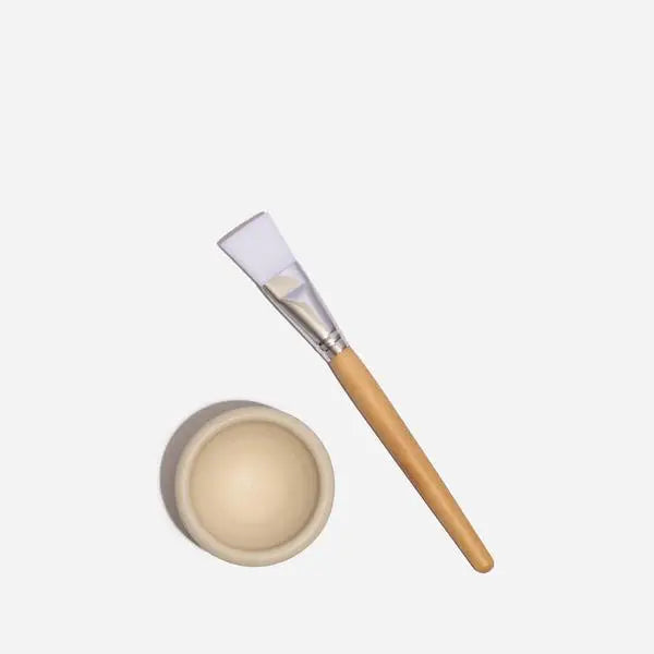 The Mask Bowl & Brush Set by Sonia's Vokalized Kreations showcases artisanal elegance with a small, beige, handmade ceramic mixing bowl and an application brush featuring a light wooden handle and white bristles on a pristine white background.