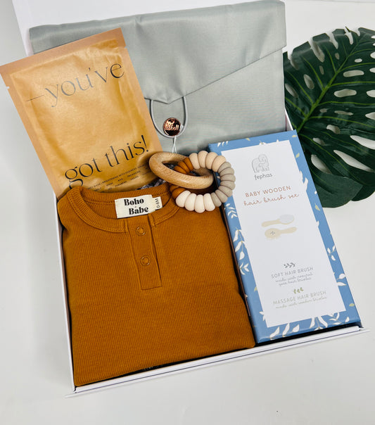 Gift box includes Sonia's Vokalized Kreations' "Mommy, Let's Cuddle!" folded brown baby outfit, a wooden teething ring, Baby Hair Brush Set in a green envelope, and an elegant card. Displayed on a white surface with a large green leaf.