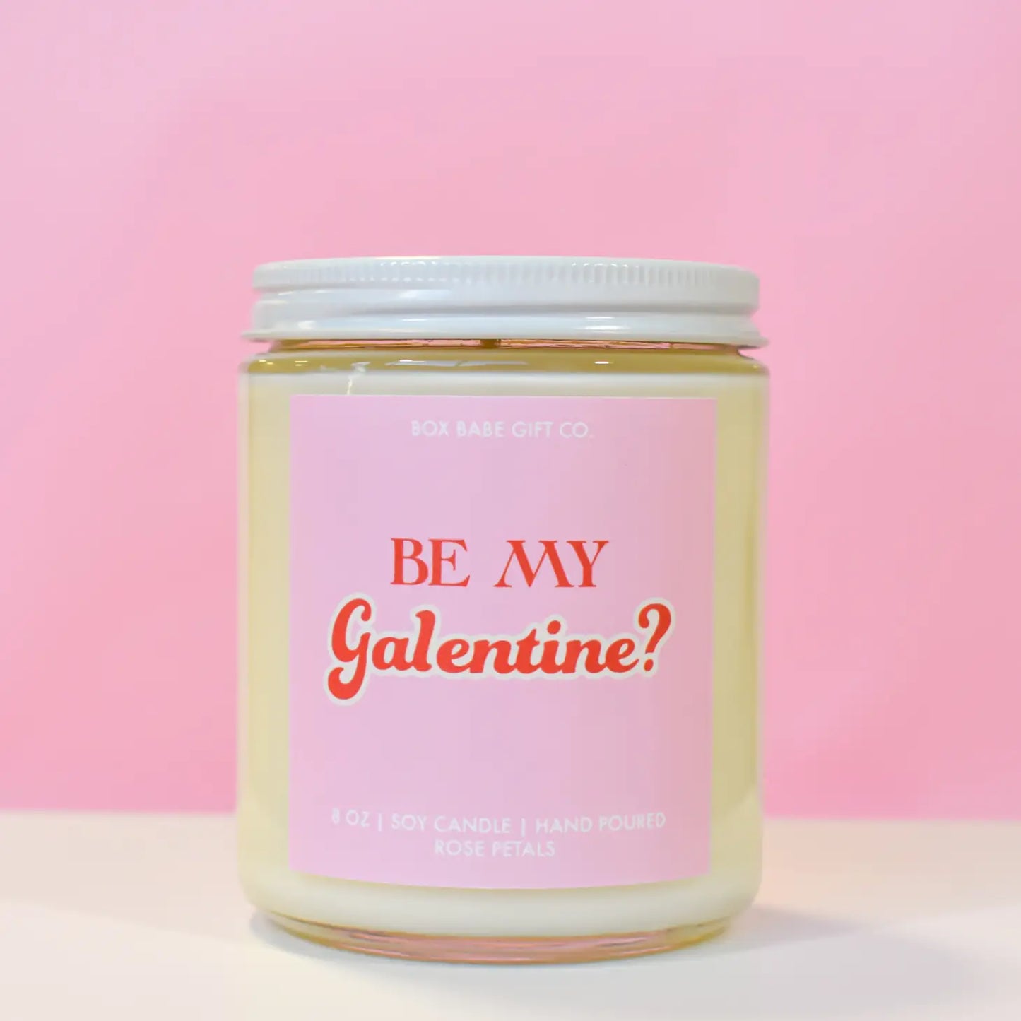 A hand-poured soy candle by Sonia's Vokalized Kreations sits on a pink background, featuring "Be My Galentine?" in red script on a glass jar with a white lid. Infused with rose petals scent, it's ideal for celebrating friendship.