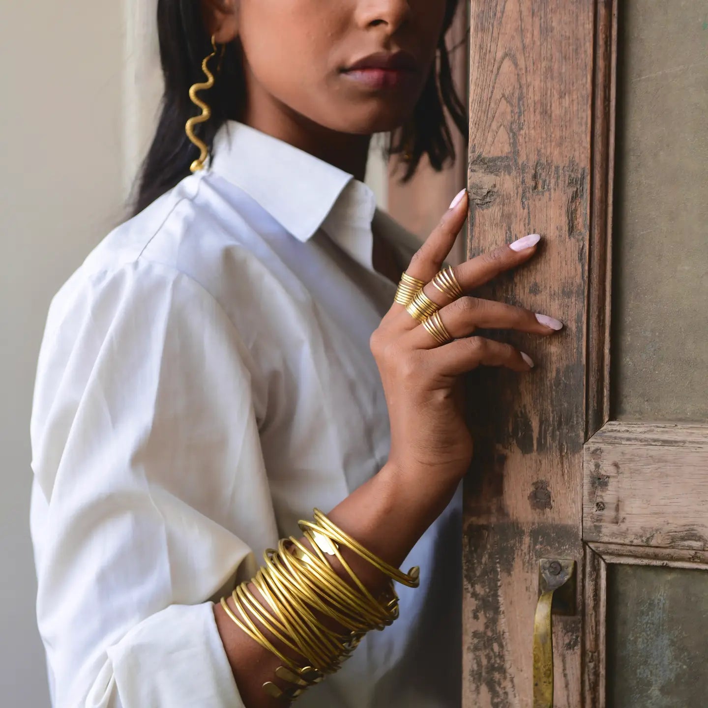 A person wearing a white shirt and gold jewelry, including Sonia's Vokalized Kreations Brass Coil Bangles, rings, and a wavy earring, stands near a wooden door, partially visible from the shoulders up.