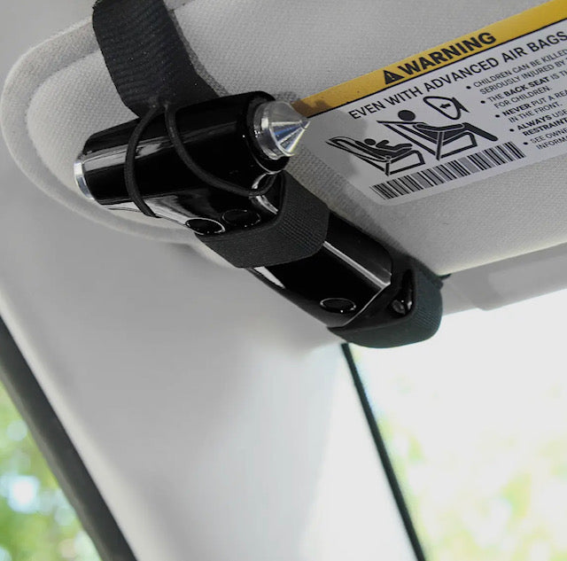 A black device, "The Glammer Hammer" by Sonia's Vokalized Kreations, is strapped to a car sun visor near an airbag warning label. It's likely a glass breaker and car escape tool.