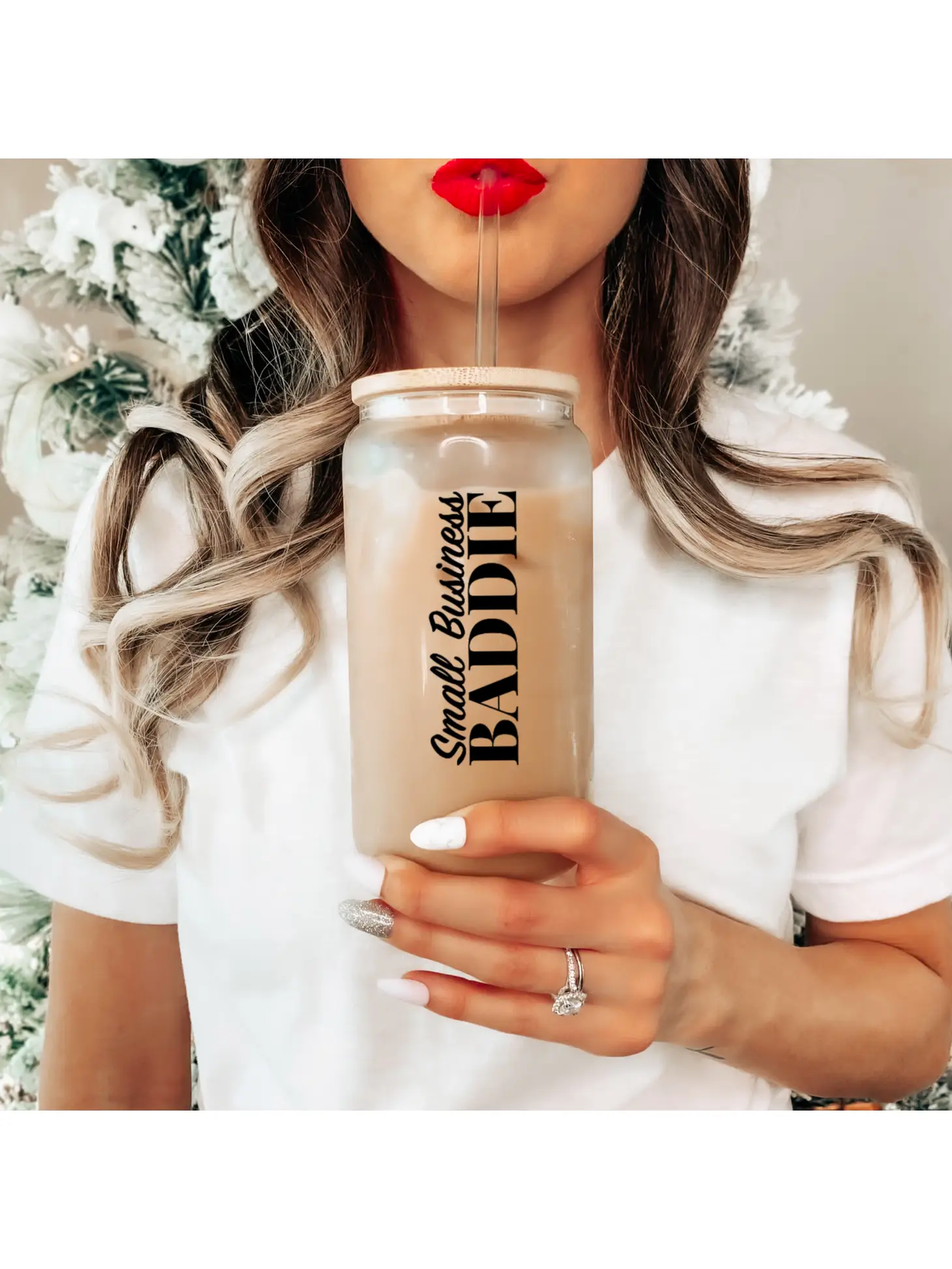 A woman in a white shirt sips an iced drink from a chic beer can glass labeled "Small Business Baddie" by Sonia's Vokalized Kreations. Set against a blurred white and green backdrop, this stylish tumbler is hand-wash only to maintain its lasting appeal. She's rocking red lipstick, embodying the baddie vibe.