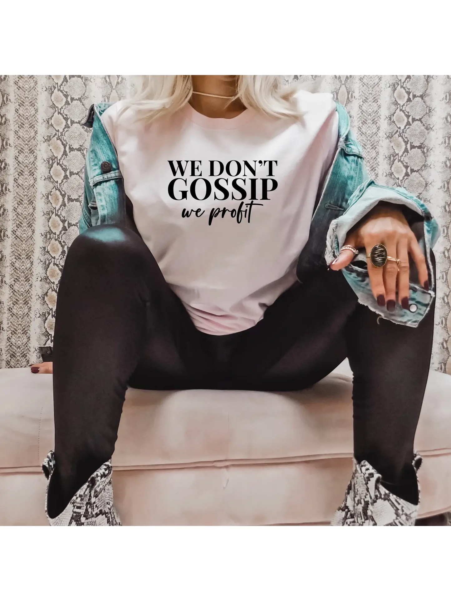 A person sits cross-legged in a "We Don't Gossip" T-shirt by Sonia's Vokalized Kreations, layered with a cotton denim jacket, black pants, and eye-catching snakeskin boots.