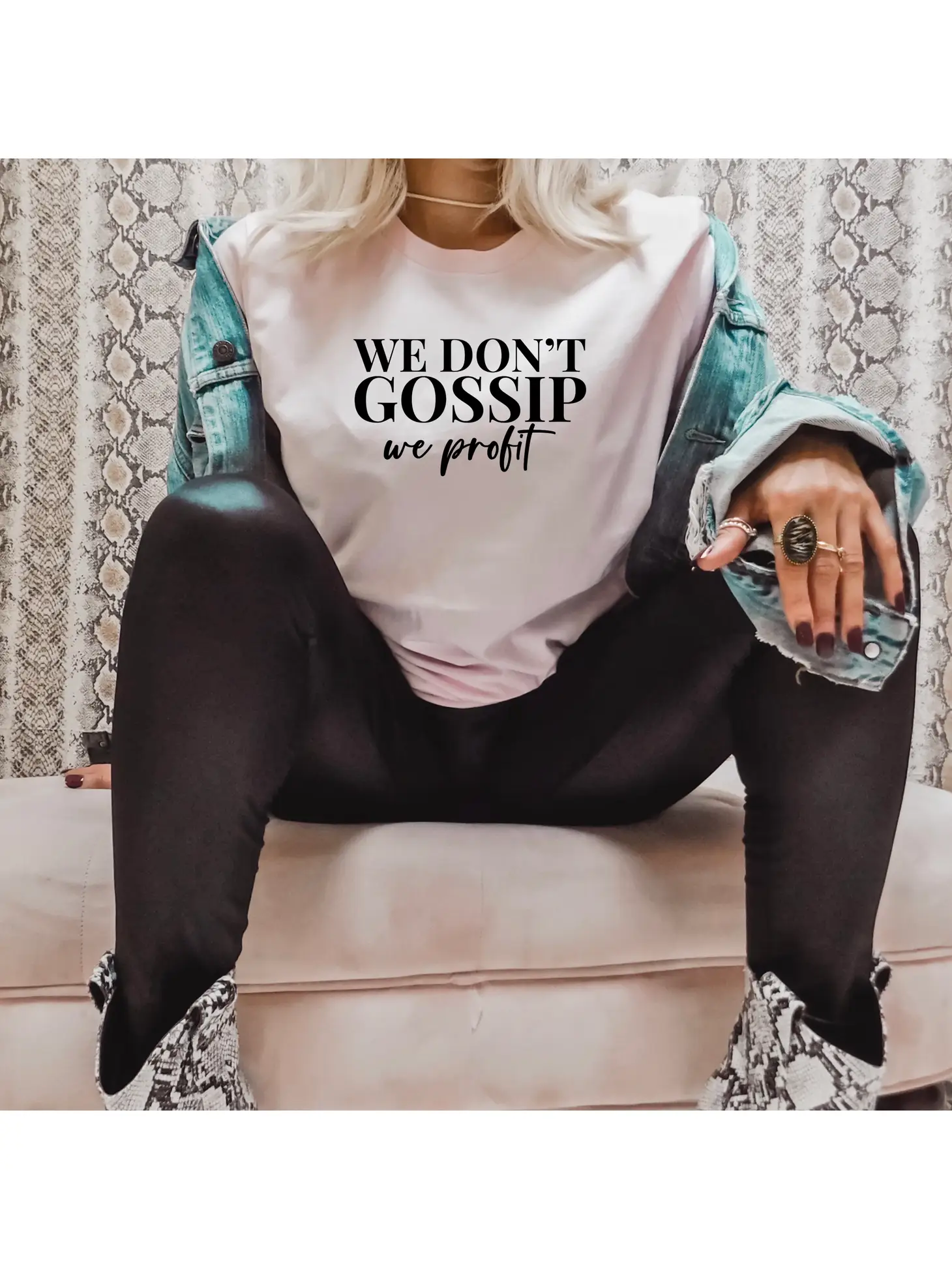 A person sits cross-legged in a "We Don't Gossip" T-shirt by Sonia's Vokalized Kreations, layered with a cotton denim jacket, black pants, and eye-catching snakeskin boots.
