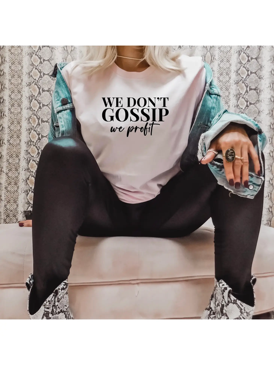 A person sits cross-legged in a "We Don't Gossip" T-shirt by Sonia's Vokalized Kreations, layered with a cotton denim jacket, black pants, and eye-catching snakeskin boots.