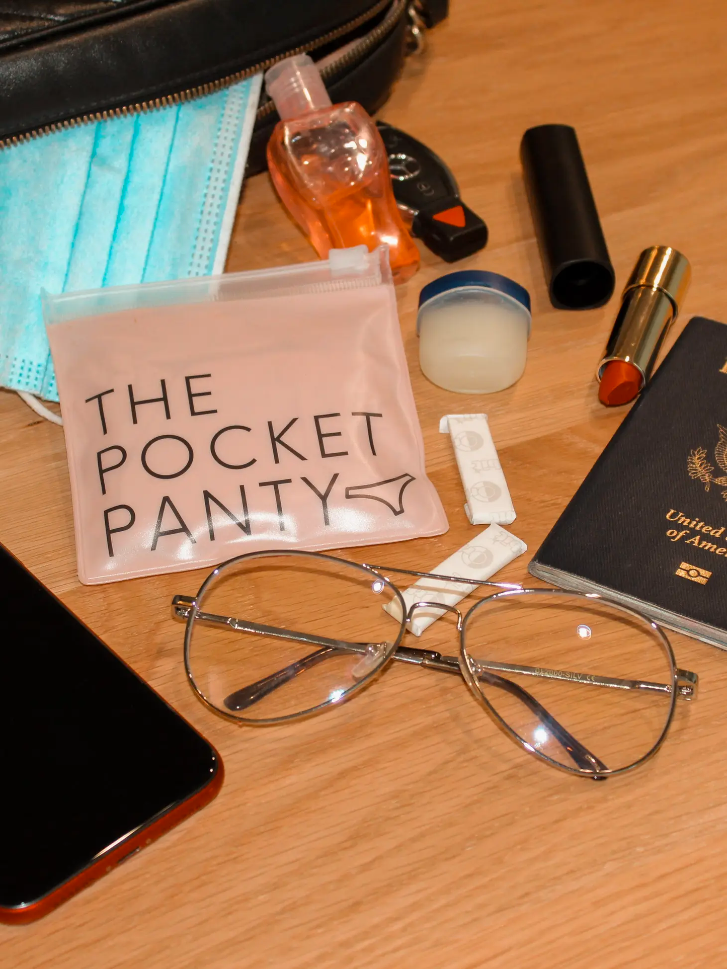 On a wooden surface: glasses, smartphone, passport, lipstick, lip balm, sanitizer, car keys, mask, tampon, and a pouch labeled "The Pocket Panty" from Sonia's Vokalized Kreations for those biodegradable panty emergencies.