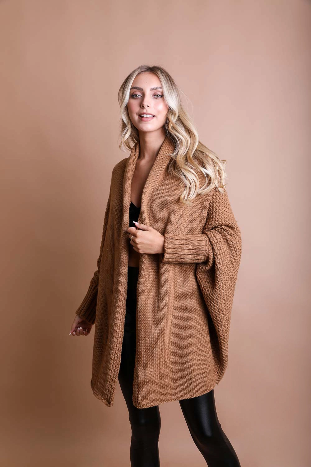 Someone wears a Bat Sleeve Knit Cardigan by Sonia's Vokalized Kreations with black pants, set against a brown background.
