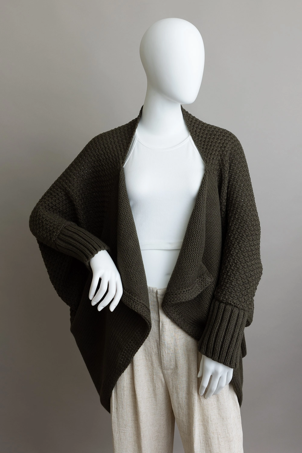 A mannequin showcases a white top, light beige pants, and Sonia's Vokalized Kreations' Bat Sleeve Knit Cardigan in dark green against a gray background, with its knit texture adding depth and interest.