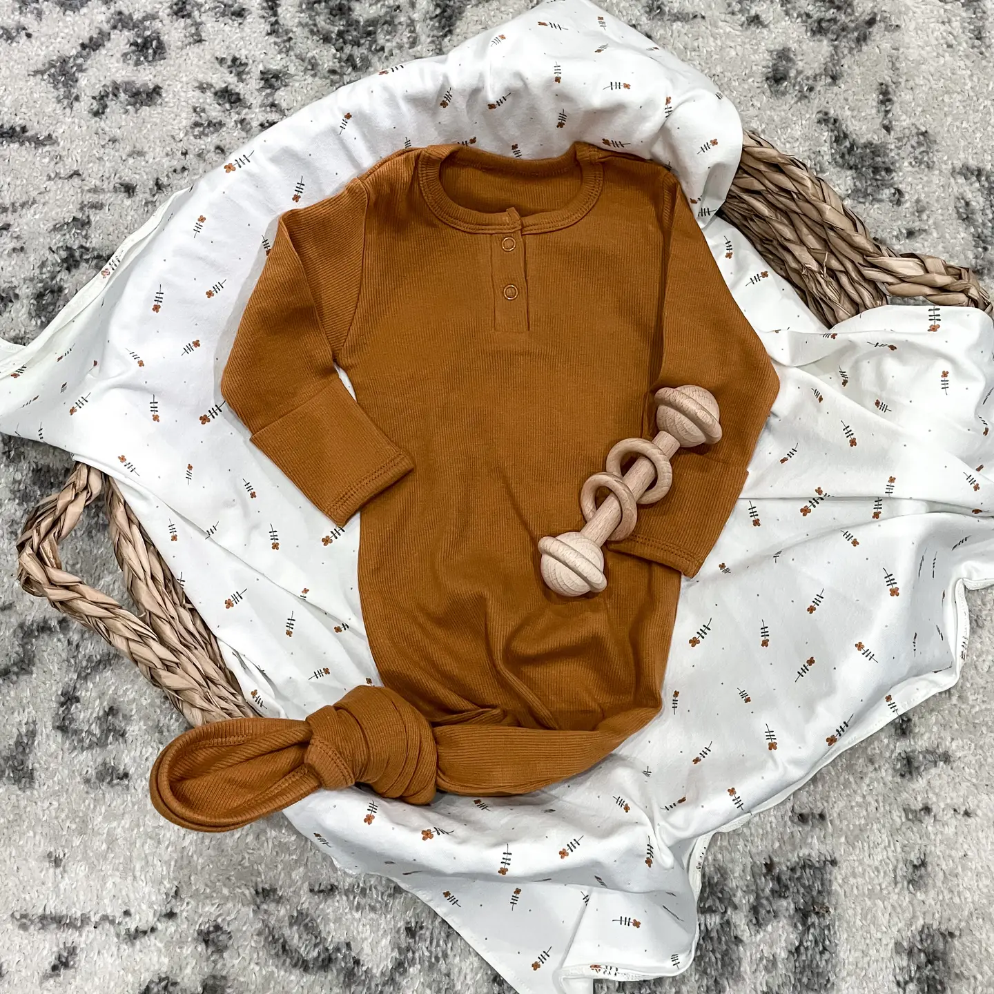 A brown baby onesie with buttons lies on a white patterned blanket in a wicker basket, alongside a beige knotted toy and the delightful "Mommy, Let's Cuddle!" print from Sonia's Vokalized Kreations.