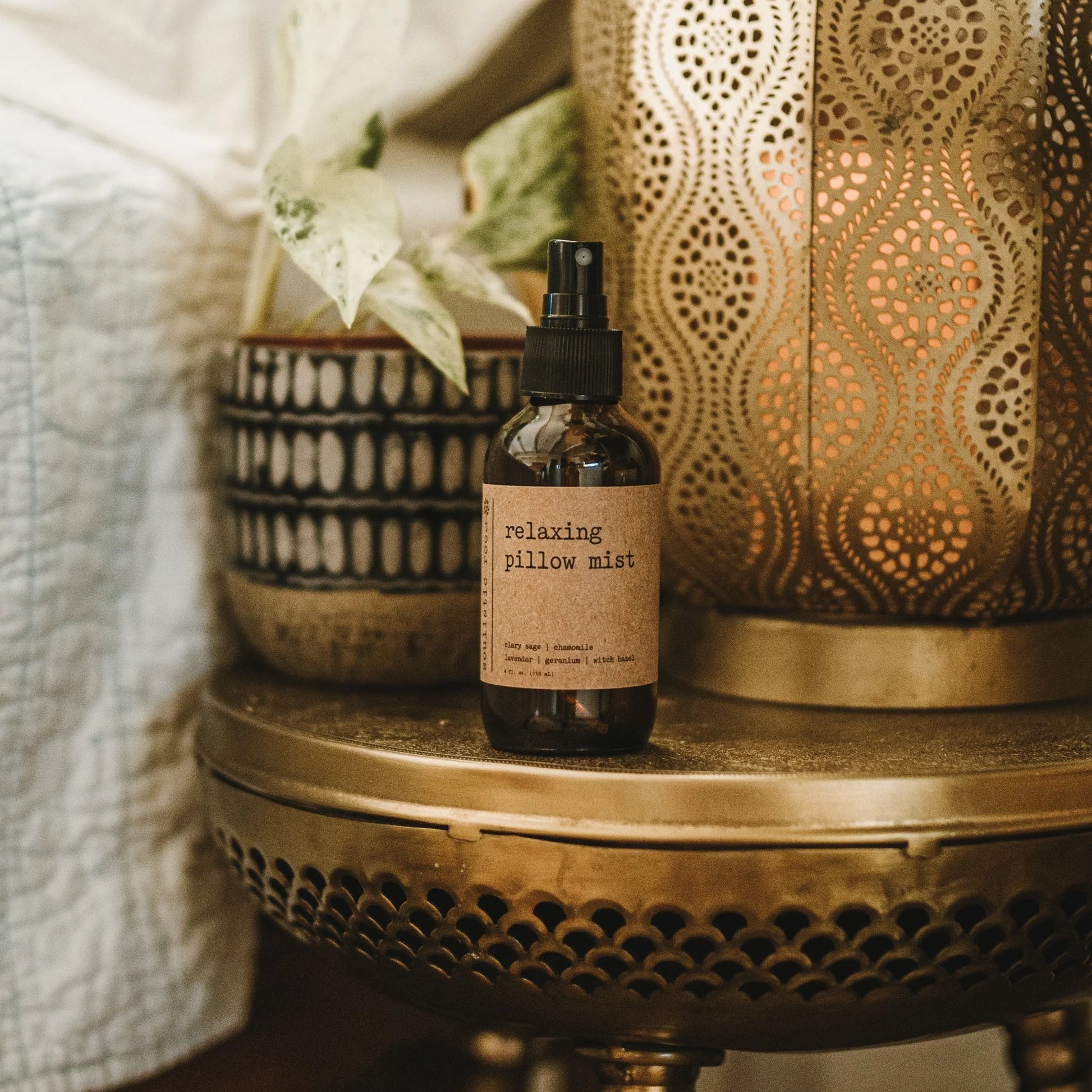 A brown glass bottle labeled "Self Less Care" by Sonia's Vokalized Kreations sits on a round, ornate brass table, creating a self-care sanctuary alongside a patterned pot with a leafy plant and a decorative metallic lantern.