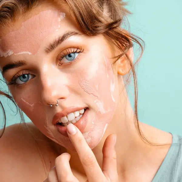 A woman softly applies Sonia's Vokalized Kreations Pink Clay Facial Mask to her face with her finger, against a calming light green backdrop.