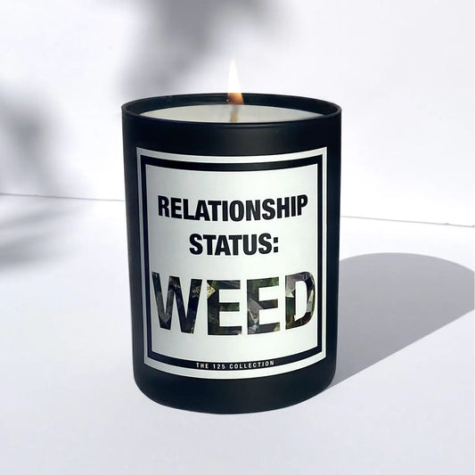 A black candle labeled "RELATIONSHIP STATUS: WEED" by Sonia's Vokalized Kreations glows, casting a shadow on a white surface. Crafted with premium fragrance oil and vegan soy wax, it's from the Relationship Status Weed collection.