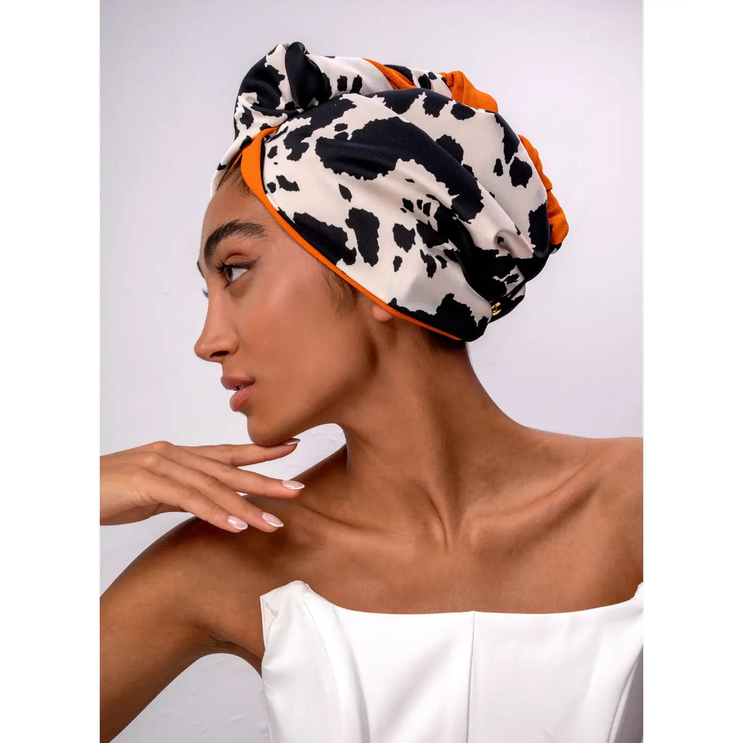 Adorned in a white strapless top and Sonia's Vokalized Kreations' "Don't Disturb My Peace" headwrap, featuring black and white patterns with orange accents, this person exudes serenity while glancing sideways against a simple backdrop.