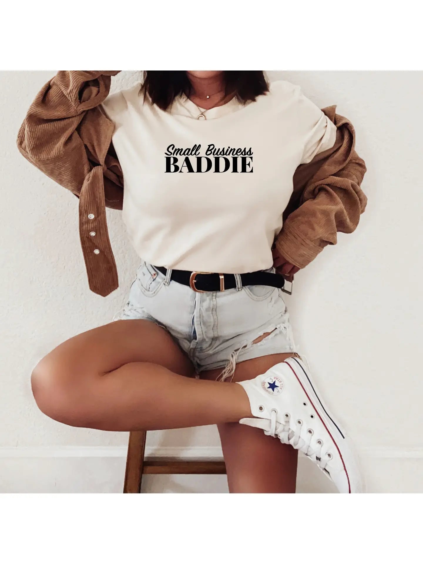 An entrepreneurial woman epitomizes the Boss Chic Essentials ethos by sonia's-vokalized-kreations, donning a "Small Business Baddie" tee with denim shorts and white sneakers. She stands confidently with one foot on a wooden chair, an unbuttoned brown jacket draping her shoulders.