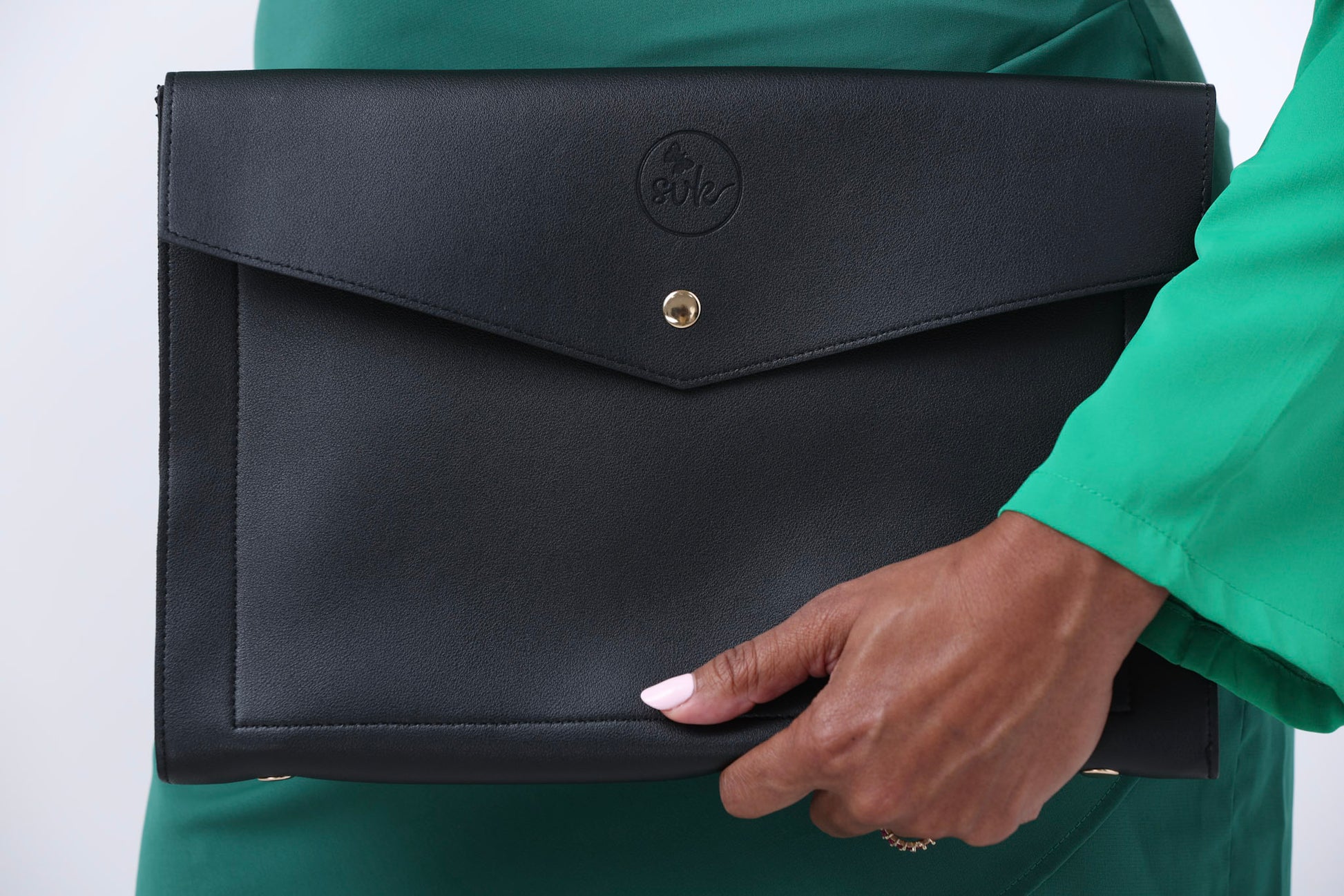A person in a green outfit holds the Harlem clutch by Sonia's Vokalized Kreations, an envelope-style piece with a round logo and gold button closure, exuding timeless elegance.