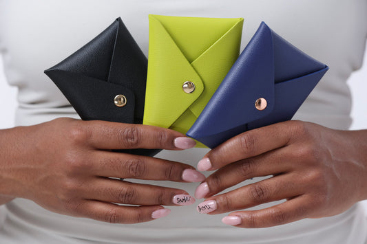 A person holds three Brooklyn Wallet 2.0s from Sonia's Vokalized Kreations—black, lime green, and blue—with compact designs. Their nails read "SAVE" and "SPEND," reflecting the ease of secure storage with snap button closures.